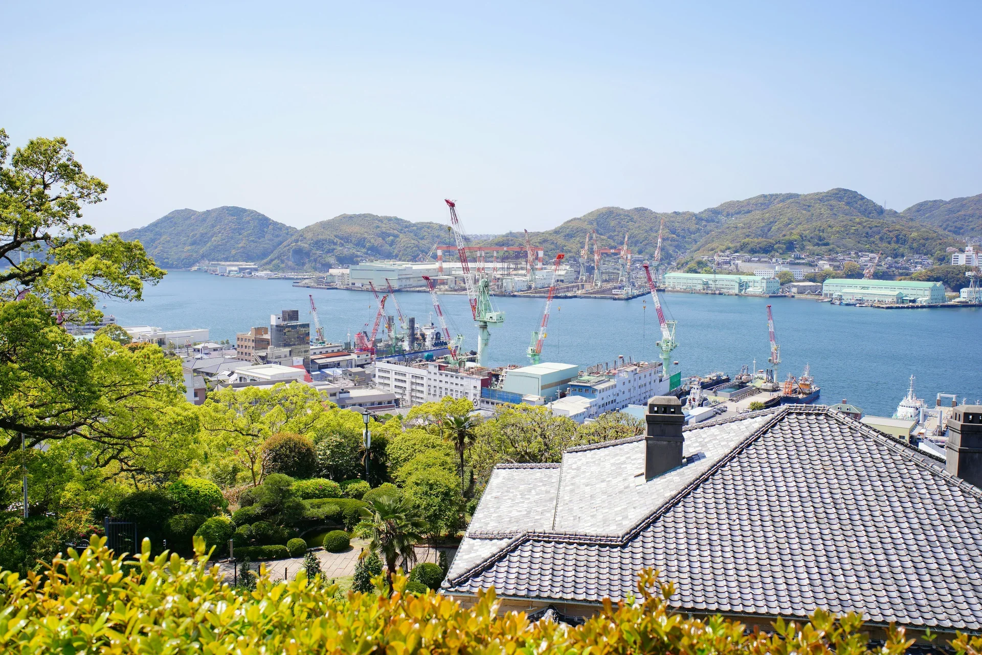 哥拉巴園俯瞰長崎港與造船廠起重機，前景是傳統瓦屋頂和園內綠樹