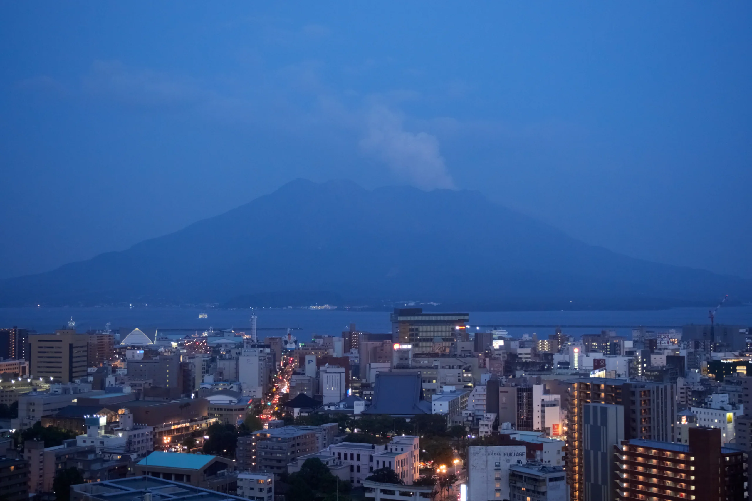 黃昏時從鹿兒島市區望向錦江灣與櫻島，遠方火山輪廓被暮色和雲氣遮住